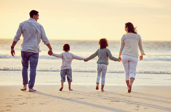 Rear view shot of a carefree family holding hands while walking on the beach at sunset. Mixed race parents and their two kids spending time together by the sea enjoying summer vacation.