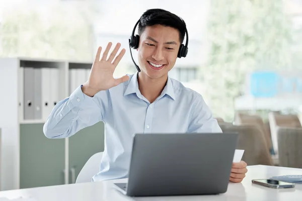 Happy asian business man in headset waving at webcam during video conference on laptop. Positive young guy in headset learning foreign language and greeting during video call in his office.