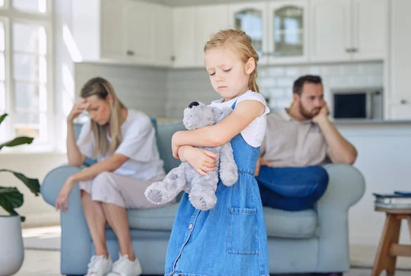 An upset little girl squeezing her teddy bear while looking sad and depressed while her parents argue in the background. Thinking about her parents breaking up or getting divorced is causing stress.