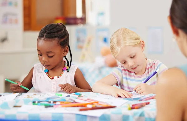 African american girl sitting at a table and colouring at pre-school or kindergarten with her caucasian classmate . Young female children using colourful pencils to draw pictures in a class at school.
