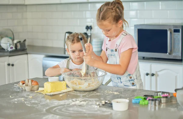 Two messy little girls baking in the kitchen at home. Caucasian focused sisters learning to make a dessert and cook. Adorable children bonding and helping each other to prepare a recipe for the house.