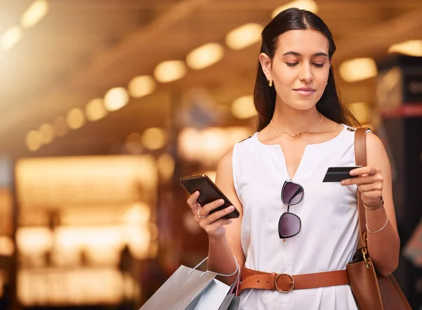 A young mixed race woman using a cell phone and bank card while carrying bags during a shopping spree. Young brunette woman purchasing items online with her smartphone and credit card in a shopping