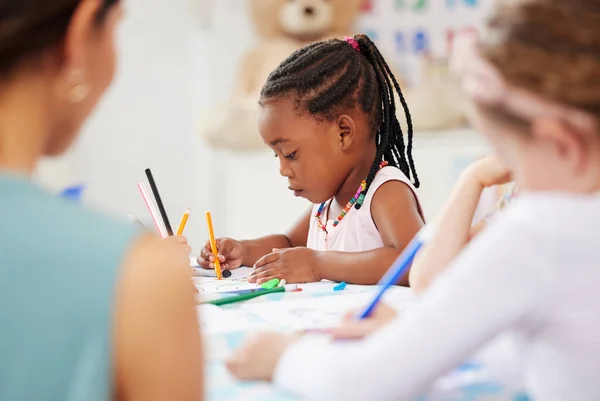 African american girl sitting at a table and colouring at pre-school or kindergarten with her fellow students. Young female child using colourful pencils to draw pictures in a classroom at her school.