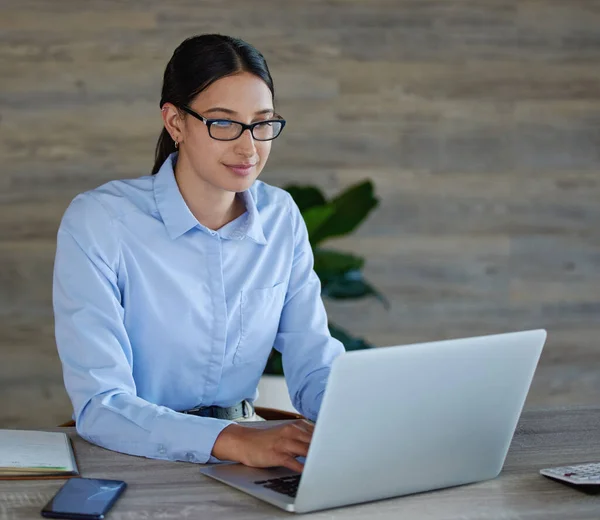 Young mixed race businesswoman wearing glasses sitting and working on a laptop alone in an office at work. One happy hispanic woman typing an email on a laptop working at desk at work.