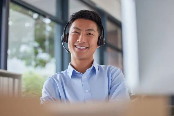 Portrait of a professional asian man smiling and wearing a headset while sitting at his desk in an office job. Happy Asian call centre agent sitting at his desk in an office.