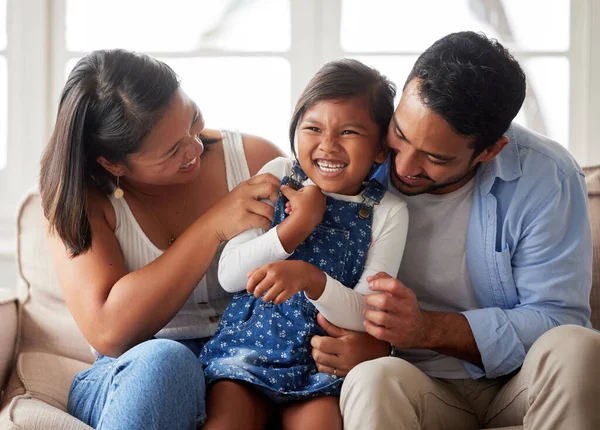 Positive family shot of a young ethnic couple smiling and laughing while bonding with their daughter on the sofa at home.