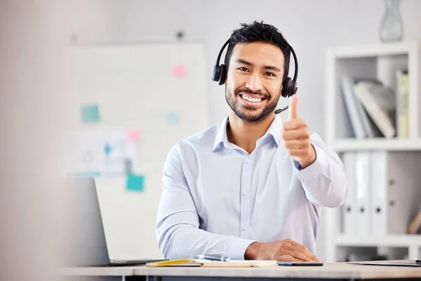 Young happy handsome mixed race male call center agent showing a thumbs up wearing a headset sitting at a desk in an office alone at work. Pleased hispanic customer service agent answering calls.