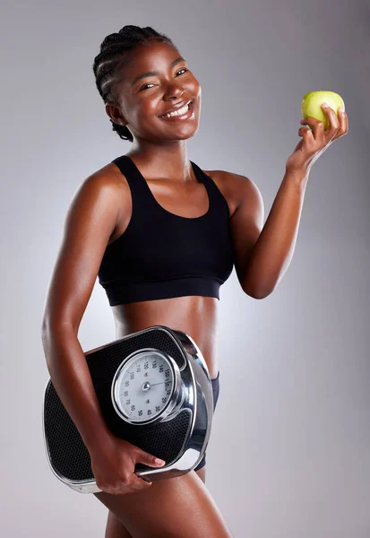 Diet and exercise can help keep you looking and feeling youthful. Studio portrait of a sporty young woman eating an apple while holding a scale against a grey background