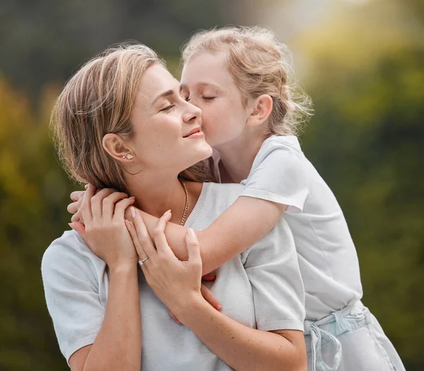 Happy and content young mother hugging and embracing her little daughter outside in the garden. Caring little girl standing while kissing her mother on the cheek and giving her a hug at a park.