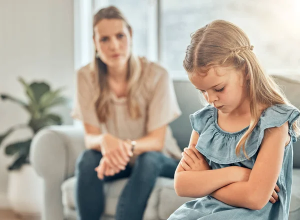 Sad little caucasian girl sitting in a living room with her arms crossed and feeling depressed while her mother watches annoyed. Young cute daughter feeling upset after disappointing a parent at home.