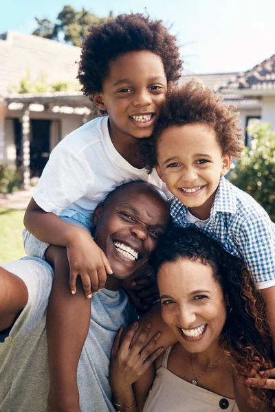 Happy african american family of four having fun while taking selfies in the sun. Carefree parents carrying sons for piggyback rides while bonding outside. Mom and dad enjoying quality time with kids.
