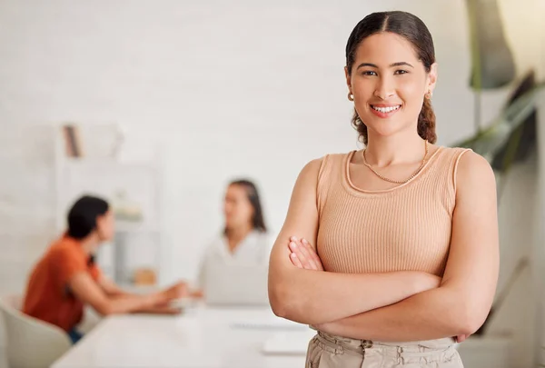 Portrait of one confident young hispanic business woman standing with arms crossed in an office with her colleagues in the background. Ambitious entrepreneur and determined leader ready for success in