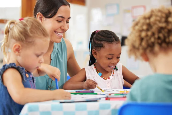 Teaching and kids looking happy while doing activity at kindergarten or pre-school. Smiling teacher sitting at table with toddlers while colouring with crayons.