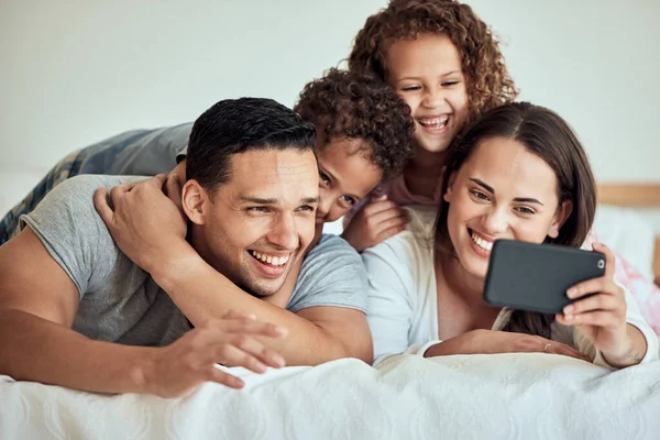 Family taking a selfie with a cellphone in bed. Happy hispanic family taking photos in bed on a smartphone. Parents making memories with their children. Excited children taking pictures.