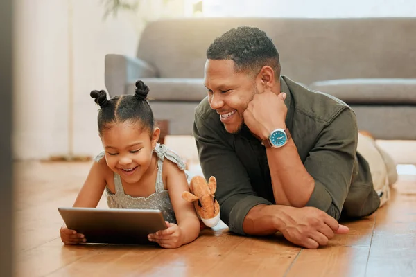 A happy mixed race family of two relaxing and lying on the lounge floor together. Loving black single parent bonding with his daughter while using a digital tablet to watch movies.