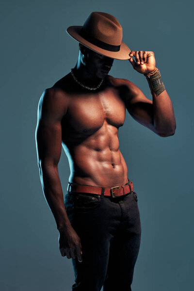 Fashionable african American model posing shirtless against blue studio background with copyspace. Sexy, serious black man with attitude showing bare six pack while wearing hat. Masculine and muscle.