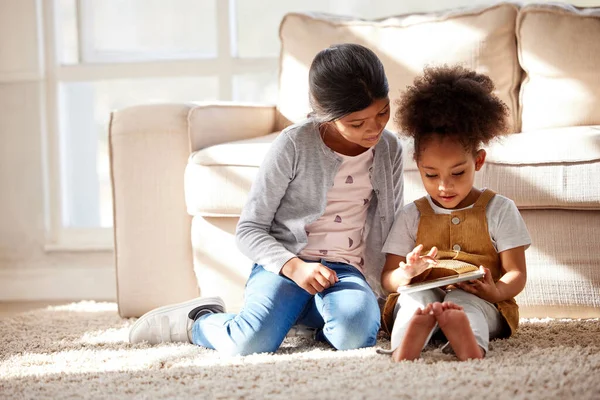 Two little mixed race sisters sitting on the floor and playing on digital tablet together at home. Hispanic female friends learning to use a digital tablet together.