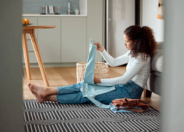 Young woman sitting on the floor folding laundry. Woman folding fresh,cleaned laundry at home. Woman cleaning her clothing and bedding. Woman busy with housework chores