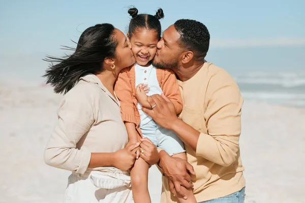 Happy mixed race family standing on the beach. Loving parents kissing adorable little daughter on the cheeks showing love and affection while enjoying beach vacation.