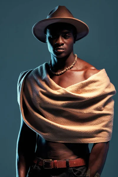 Fashionable african American model posing with wrapped scarf against blue studio background with copyspace. Sexy, serious, handsome black man with attitude wearing hat and shawl. Masculine and muscle.