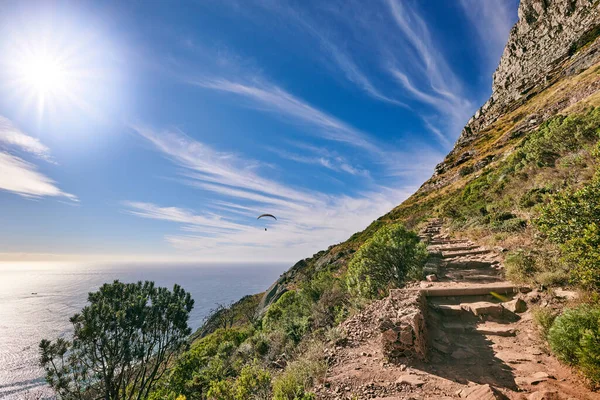 A hiking trail up a rocky mountain with green plants growing and cloudy blue sky background. The landscape of a path in a beautiful coastal trekking location near the ocean outdoors in nature.
