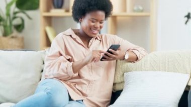 Evde telefonla mesajlaşırken gülümseyen ve gülen genç bir kadın. Neşeli bir kadın sosyal medyada arkadaşlarıyla sohbet ediyor, internette geziniyor ve kanepede dinlenirken komik internet mimleri izliyor..