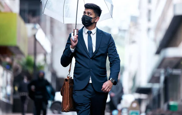 Paving his path in the new world. Portrait of a young businessman wearing a face mask and holding an umbrella on a rainy day in the city