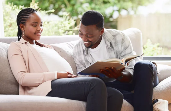 Let me read you a story. a handsome young man reading a book to pregnant wifes belly while sitting on the sofa at home