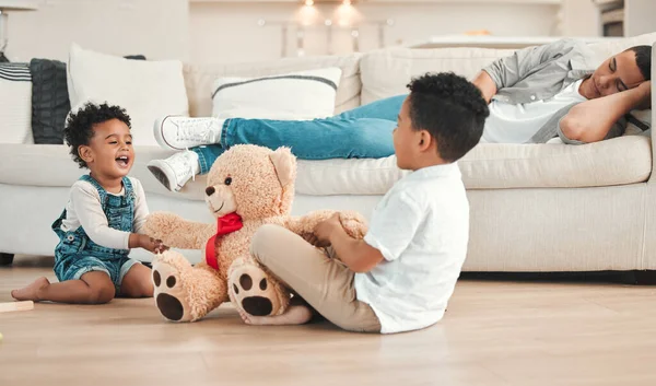Dad needs a break. two siblings fighting over a teddy on the floor while their dad sleeps on the couch at home