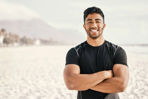 Lets go for a run. a sporty young man standing with his arms crossed while out for a workout at the beach