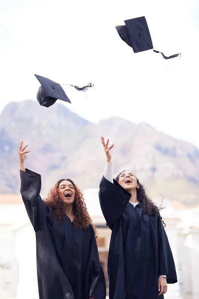 The harder you work, the higher the reward. two young women throwing their caps in the air on graduation day