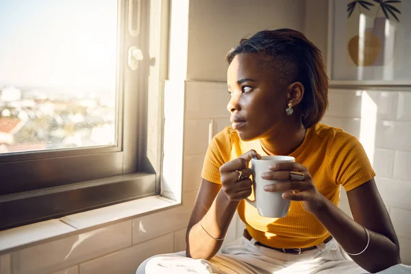A thinking and coffee drinking black woman enjoys her morning routine while gazing outside a window on a bright sunny day. Beautiful female looks thoughtfully towards city while sitting at home.