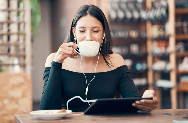 Beautiful, happy and relaxed student with tablet drinking coffee, listening to a podcast and music on earphones in a cafe. Woman online and watching distance learning education webinar in restaurant.