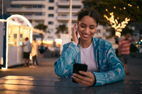 Modern, trendy and happy woman texting on a phone, listening to music and smiling while browsing social media at at an outdoor cafe. Young female enjoying night life in the city while chatting online.