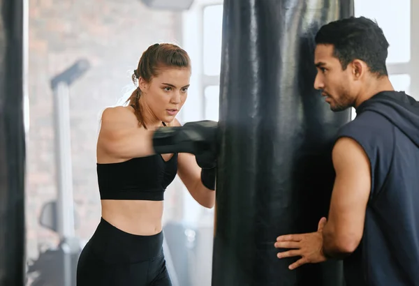Fit female learning boxing on a punching bag and getting fitness training advice from her personal trainer at a gym studio. Strong boxer with slim body practicing for a fight with her fighting coach.
