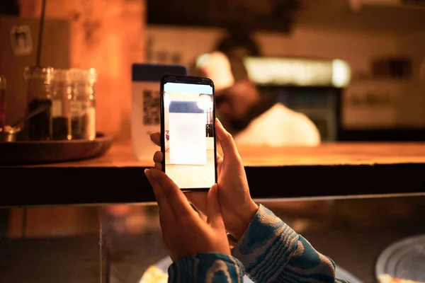 Person with phone camera scanning QR bar code for online menu at a modern restaurant, shop or cafe at night. Hand of a woman taking photo scan for website information using modern technology.