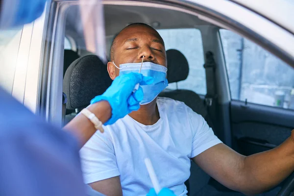 Covid testing and screening of a man driving in his car at a drive through station with medical nurse assistance. Guy getting virus treatment test while wearing a protective face mask.