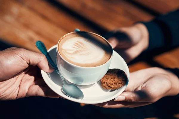Closeup of hands holding and giving a cappuccino or flat white to a customer at a cafe. Hot beverage with creative plant image in milk foam. A morning coffee drink made by a barista in a restaurant