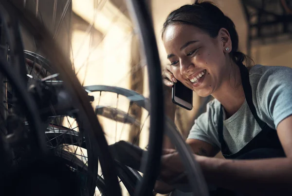 The best distraction is doing what youre good at. a young happy young woman talking on a cellphone and using a digital tablet while fixing a bike at a bicycle repair shop