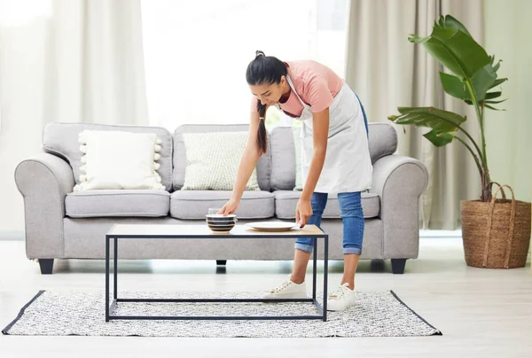 Cleaning up the mess from last nights binge-watching session. a young woman removing dirty dishes from a coffee table in the living room at home