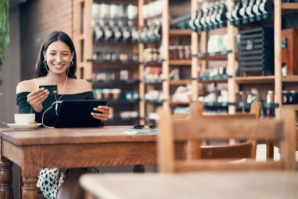 Online shopping with a credit card at a cafe by a trendy woman browsing on a tablet with earphones and paying for her bill at a coffee shop. Happy woman enjoying a break and downloading a finance app.