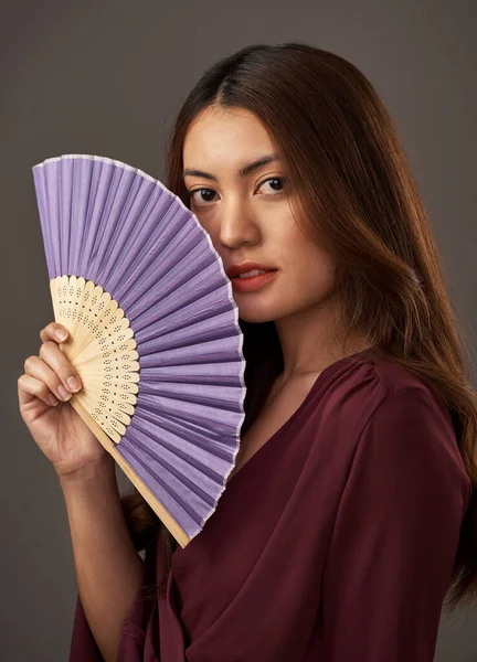 Fan the flames of beauty. Cropped portrait of an attractive young woman posing with a fan in studio against a grey background