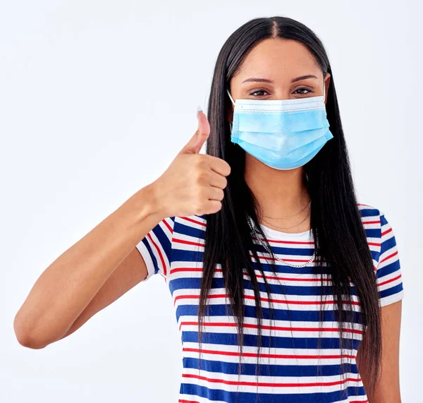Keeping a positive mindset in the pandemic. Studio portrait of a young woman wearing a face mask and showing thumbs up against a white background