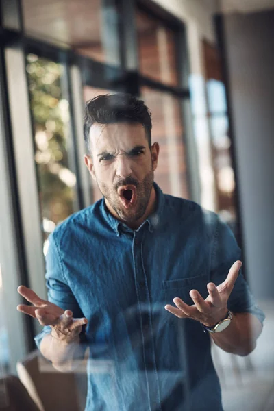 Angry, stressed and frustrated man unhappy and feeling bad about work problems while standing in a modern office alone. Aggressive, annoyed and upset young male filled with emotions screaming loud.