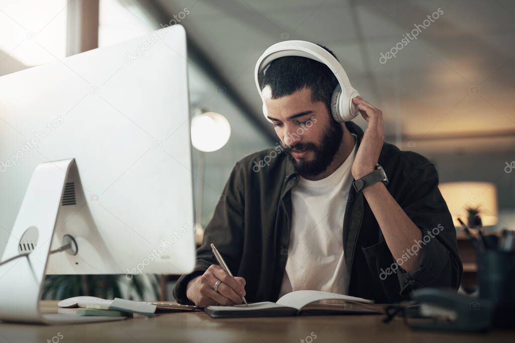 Good music keeps the creative juices flowing. a young businessman writing in a notebook and using a computer with headphones during a late night at work