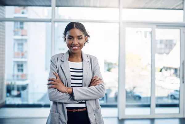 Confident, proud and professional young black business woman folding her arms and smiling in a modern office. Portrait of African American leader looking powerful, successful standing in power stance.