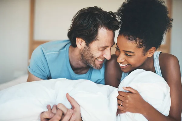 Happy, loving and funny interracial couple smiling and laughing while lying in bed and sharing a tender moment. Carefree husband and wife relaxing and having fun while bonding in the bedroom.