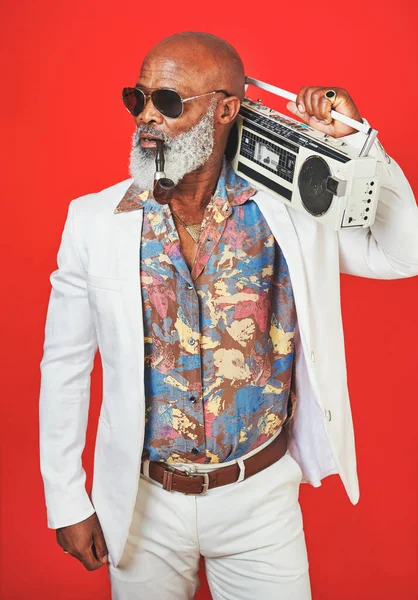 Ill take you back, way back. Studio shot of a senior man wearing vintage clothing while posing with a boombox against a red background