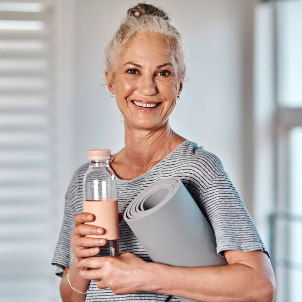 I have everything I need to get started with my workout. Portrait of a cheerful mature woman holding a bottle of water and yoga mat ready to start her morning session of yoga inside of a studio