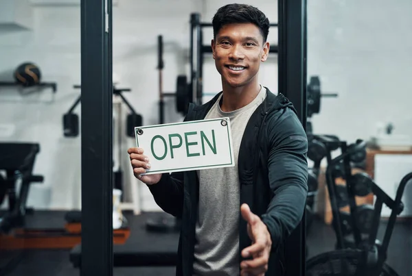 Let me welcome you to our fitness centre. Cropped portrait of a handsome young male fitness instructor extending his hand for a handshake while holding up a sign that says OPEN in a gym